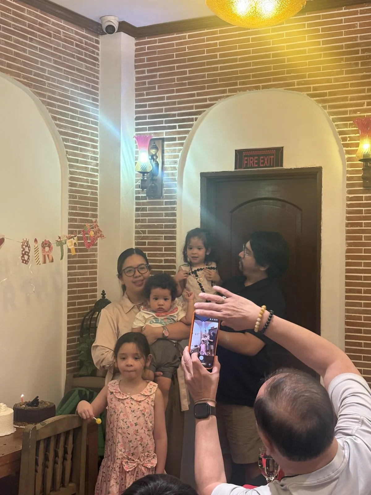 Carla holding Ottoman and Mikko holding Bebu, with Lola beside them by the birthday banner while a relative takes their photo