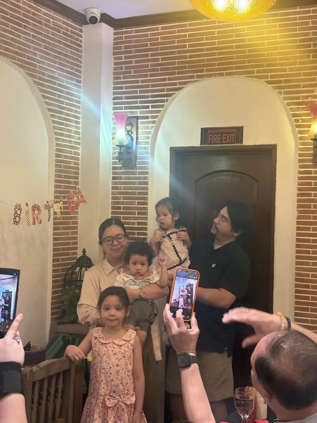 Carla holding Ottoman and Mikko holding Bebu, with Lola, smiling by the fire exit door as relatives take photos at Mr Kebab, West Ave