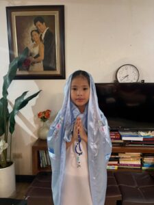 Chloe dressed as Mama Mary in a light-blue veil, holding a rosary