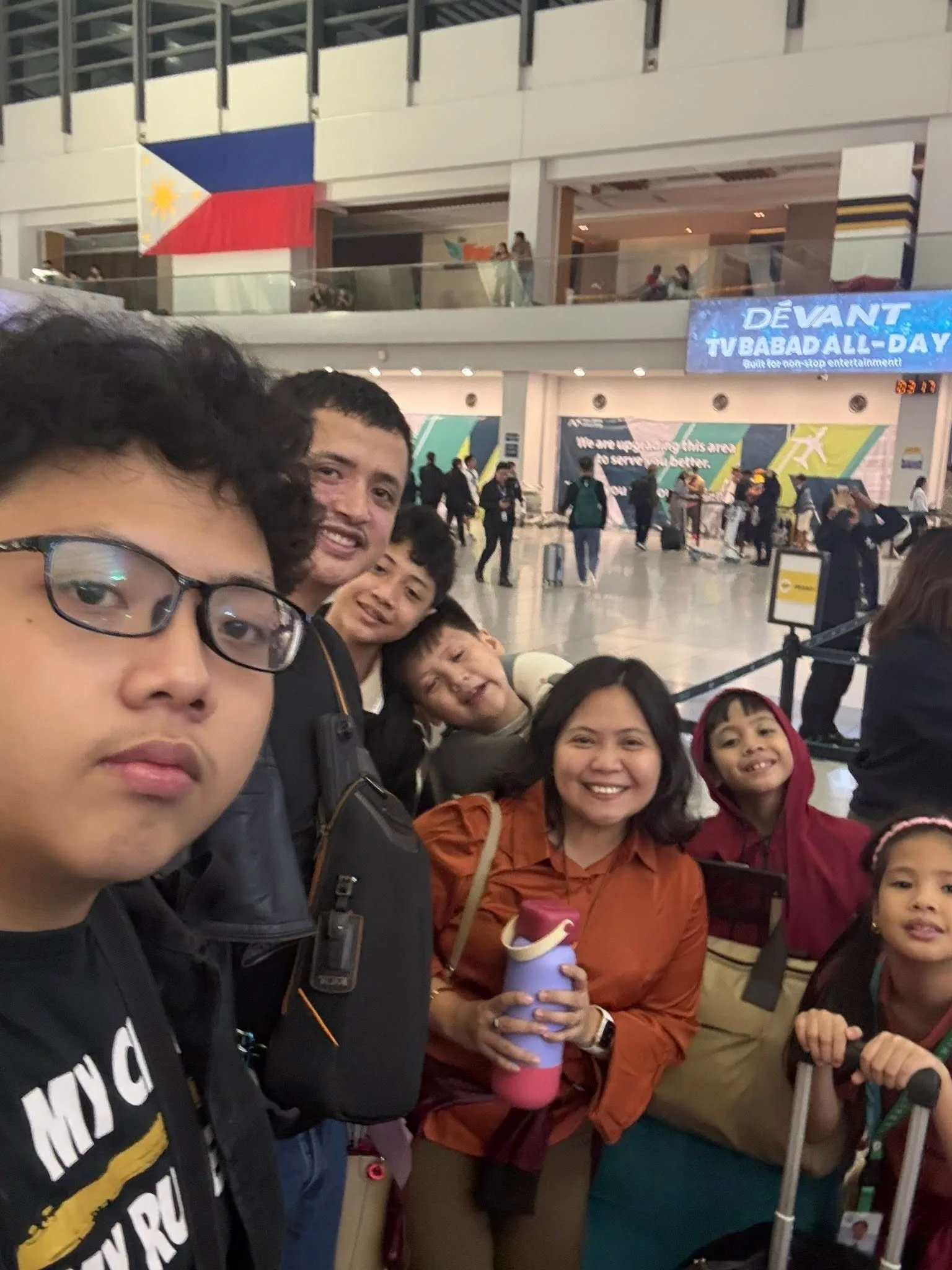 Family at NAIA Terminal 3 departure area with the Philippine flag behind, ready for Singapore