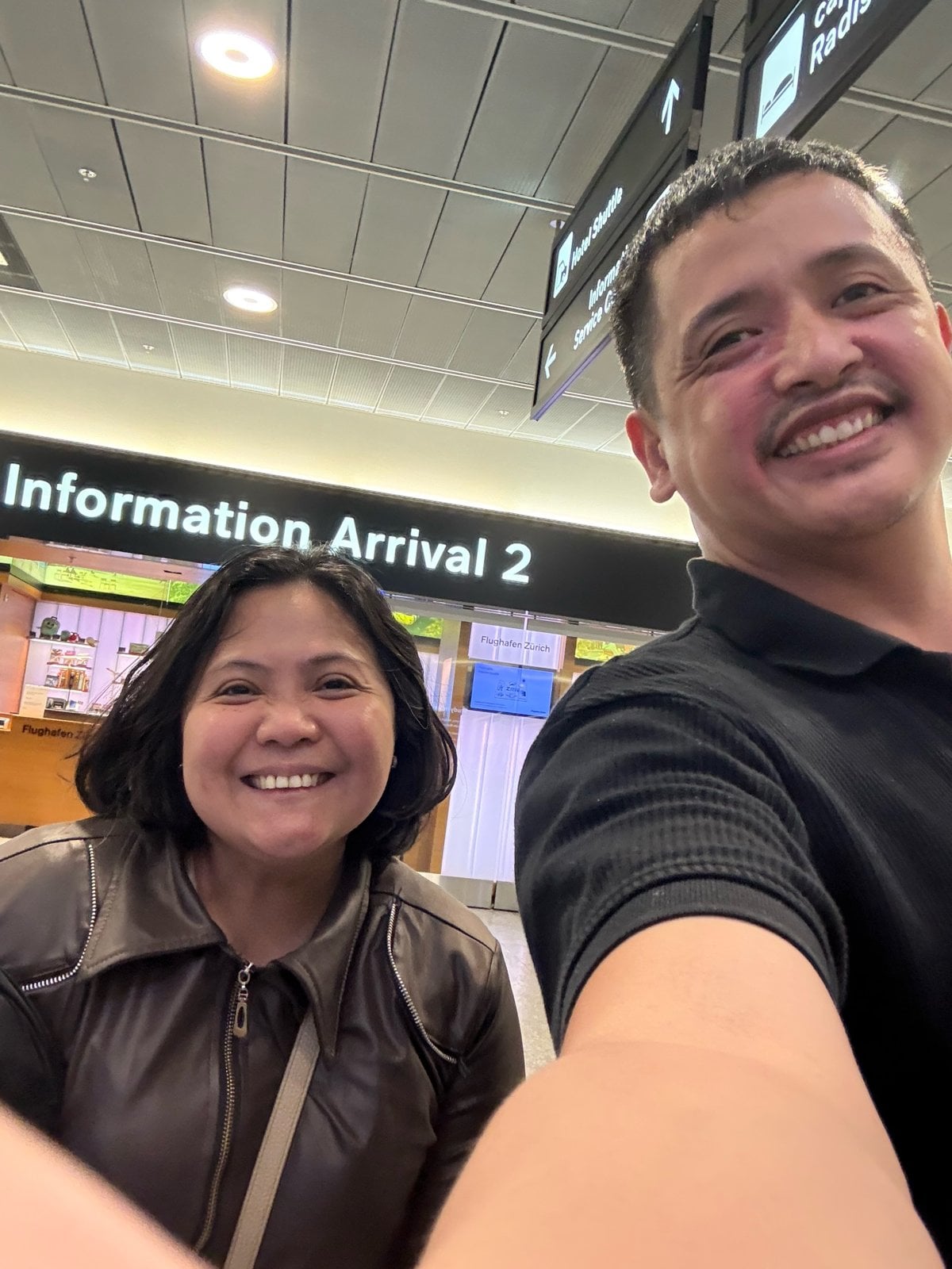 Paolo and Rosed smiling under the Information Arrival 2 sign at Zurich Airport