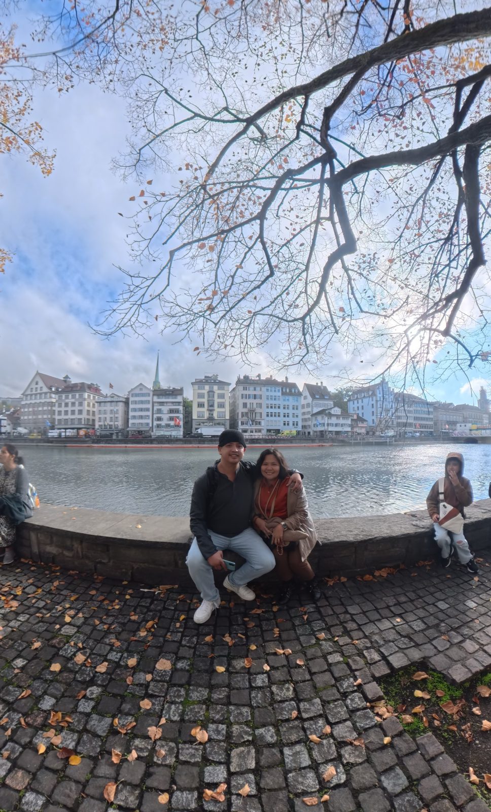 Wide shot of Paolo and Dimple on the Limmat river wall under autumn branches